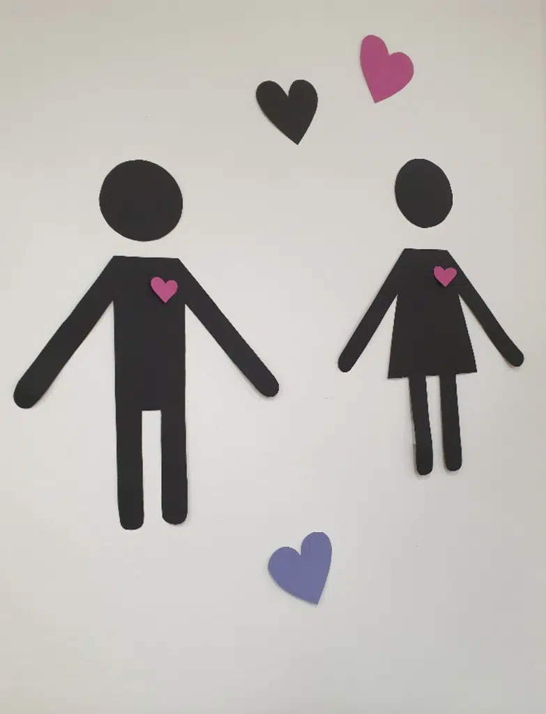 Zwei Menschen (eine Frau, ein Mann) sind als schwarze Piktogramme an einer weißen Wand aufgehängt. Dazu drei Herzen, ebenfalls im Piktogrammstil