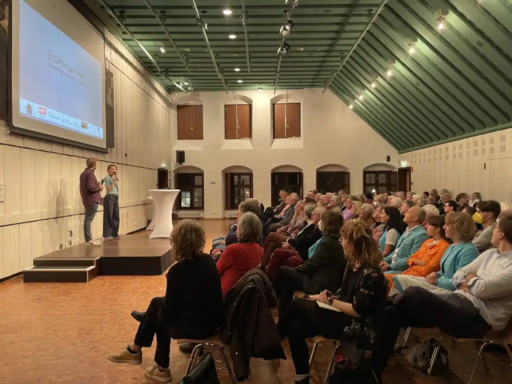 Blick in den Saal des Münchner Stadtmuseums. Zwei Moderator*innen stehen auf der Bühne, viele Zuschauer sind im Publikum