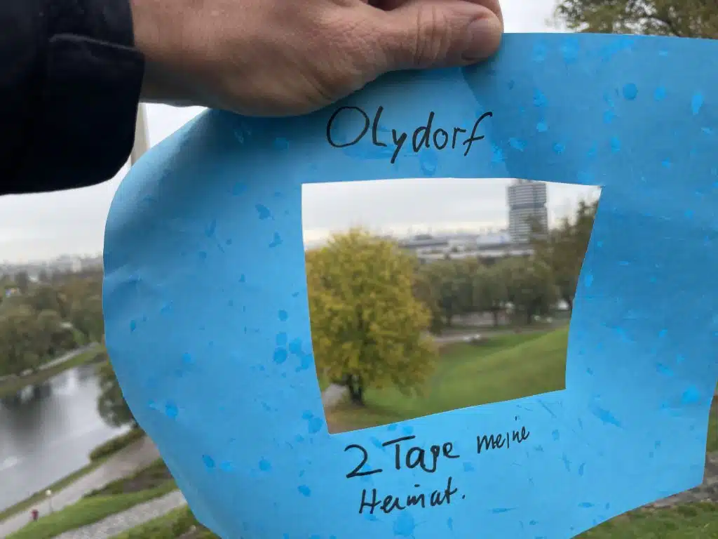 Ein Papierrahmen, in dessen Mitte der Hintergrund (Olympiapark) zu sehen ist. Auf dem Rahmen steht handschriftlich geschrieben: Olydorf, 2 Tage meine Heimat