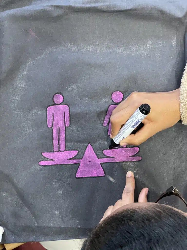 Eine Schülerin zeichnet mit einem pinken Filzstift ein Piktogramm auf ein Stück Stoff: Zu sehen ist eine Waage, links steht ein Mann und rechts eine Frau.